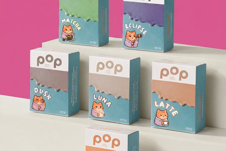 Six boxes of 'pop' color lenses arranged on white steps against a pink background. The boxes feature different colors, each with a cute cartoon cat illustration and the flavor name like 'MATCHA', 'ECLIPSE', 'DUSK', 'LUNA', 'LATTE', and 'HALO'.